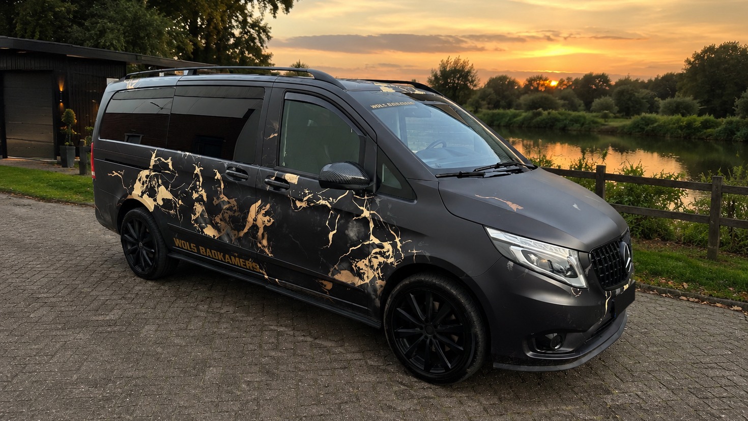 De matzwarte bedrijfsbus van Wols Badkamers met gouden marmerlook belettering, bij een rivier tijdens zonsondergang